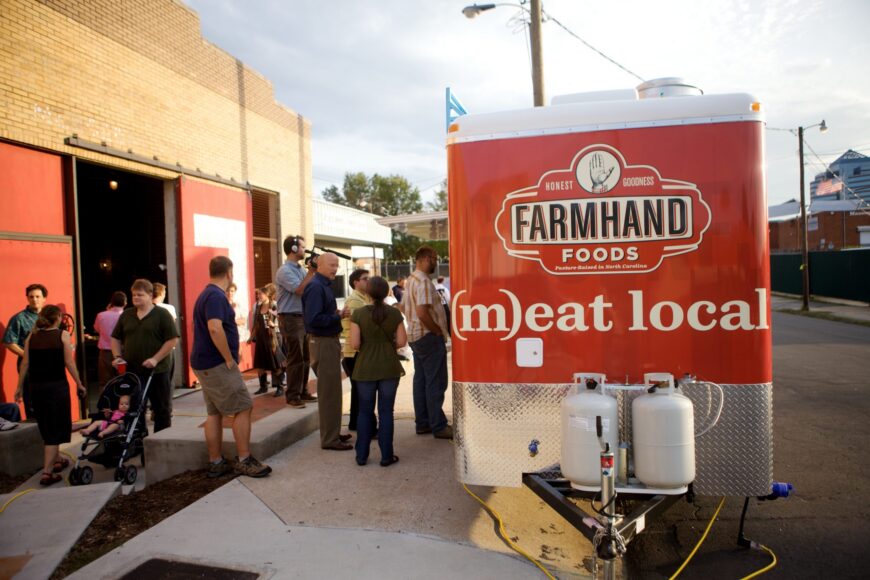The back of the red Farmhand Foods Sausage Wagon, showing the original hand logo and the words (m)eat local, with a crowd gathered outside a brick building in Durham, North Carolina.